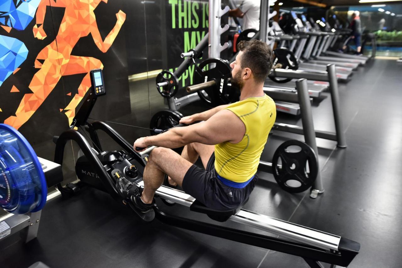 ดอสโซ่ ดอสซี่ โฮเต็ล แอน์ สปา ดาวน์ทาวน์ Hotel อิสตันบูล ภายนอก รูปภาพ The photo shows a man exercising on a rowing machine in a gym. He is wearing a yellow tank top and black shorts. The background features an exercise-themed mural with colorful graphics of a person in motion. Various gym equipment, including treadmill