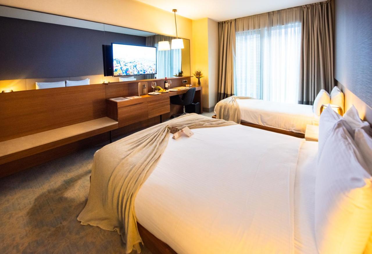 ดอสโซ่ ดอสซี่ โฮเต็ล แอน์ สปา ดาวน์ทาวน์ Hotel อิสตันบูล ภายนอก รูปภาพ The photo shows a modern hotel room featuring two beds with white linens and a decorative throw blanket. There's a wooden desk and a chair alongside a large flat-screen TV mounted on the wall. The room has large windows with curtains, allowing natura
