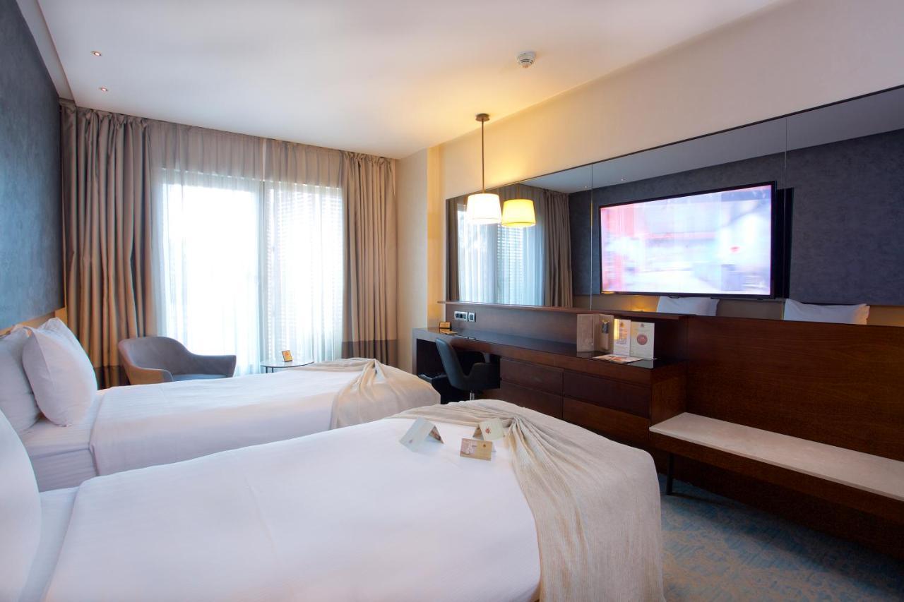 ดอสโซ่ ดอสซี่ โฮเต็ล แอน์ สปา ดาวน์ทาวน์ Hotel อิสตันบูล ภายนอก รูปภาพ The photo shows a hotel room featuring two twin beds with white linens. There is a large window with curtains that allows natural light to enter the room. A wooden cabinet is present, along with a flat-screen television mounted on the wall. Additiona