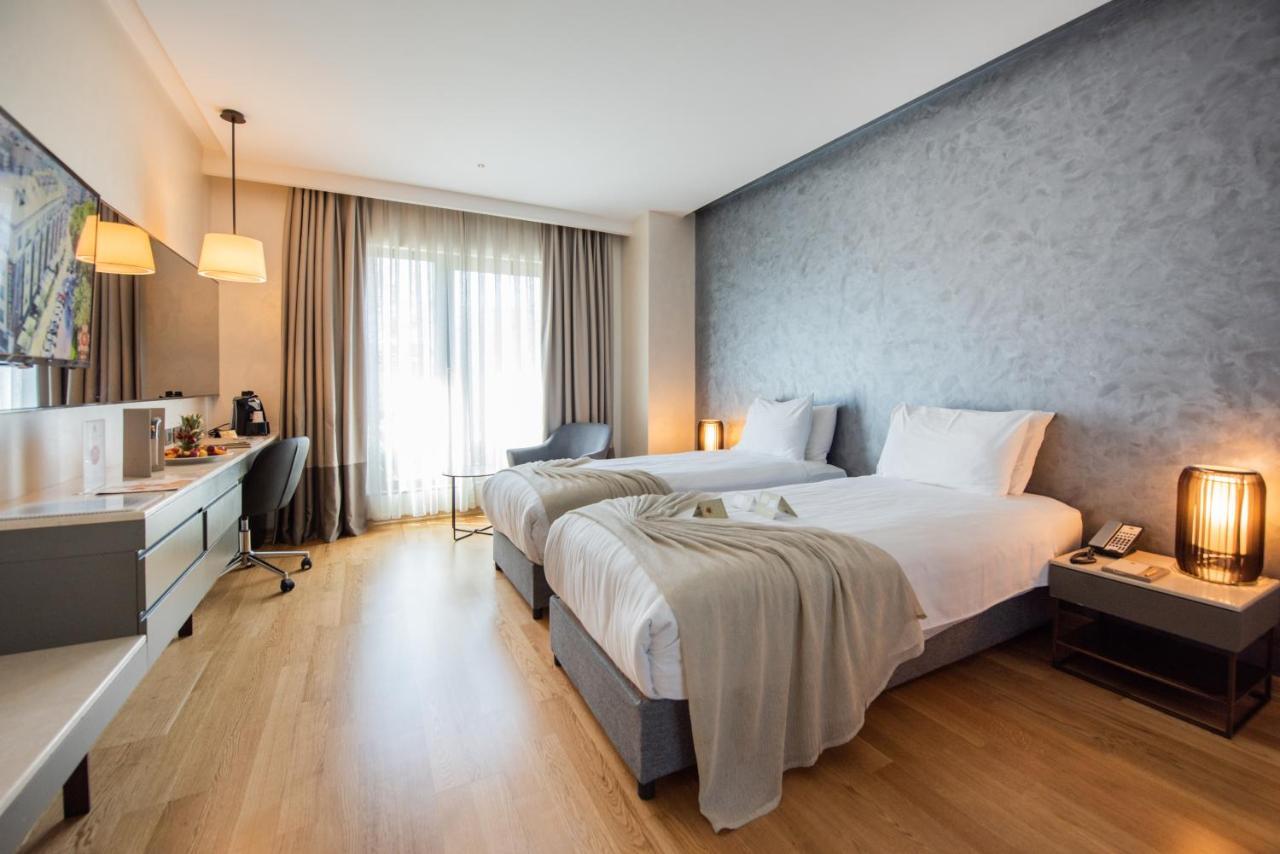 ดอสโซ่ ดอสซี่ โฮเต็ล แอน์ สปา ดาวน์ทาวน์ Hotel อิสตันบูล ภายนอก รูปภาพ The photo shows a modern hotel room featuring two single beds with white linens. The room has a muted color palette, including a gray wall and light wooden flooring. Large windows allow natural light to flood the space, and there are sheer curtains f