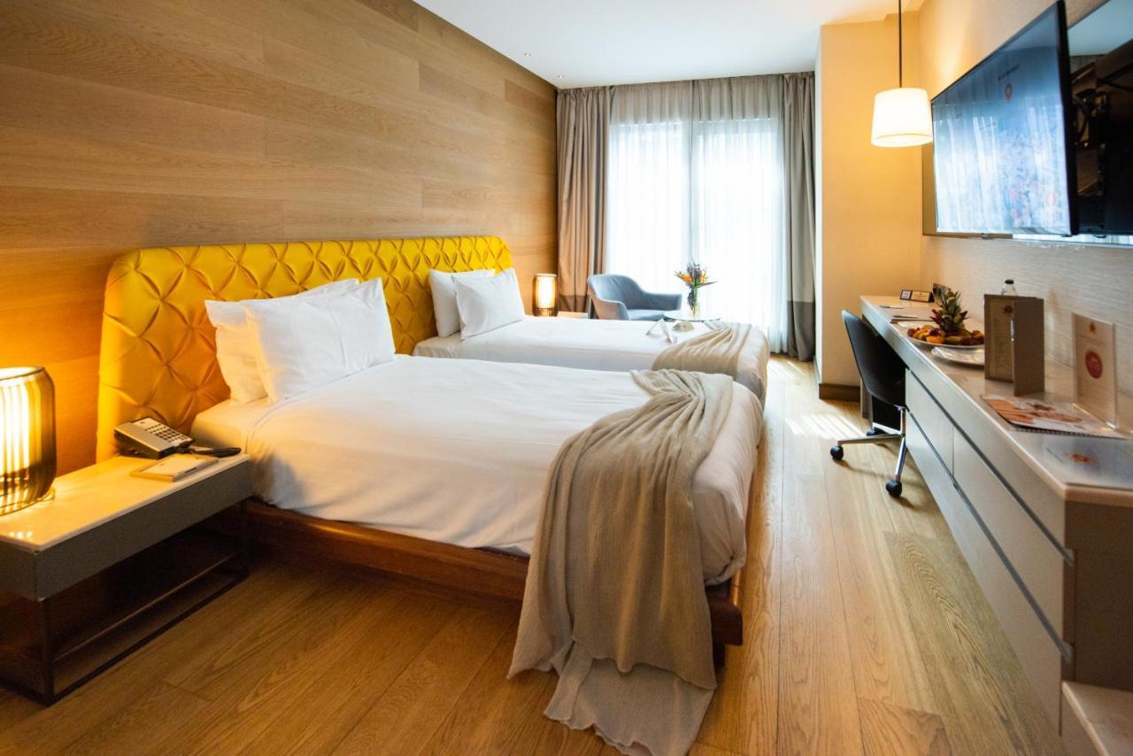 ดอสโซ่ ดอสซี่ โฮเต็ล แอน์ สปา ดาวน์ทาวน์ Hotel อิสตันบูล ภายนอก รูปภาพ The photo shows a stylish hotel room featuring two beds with white linens and a unique yellow headboard. The room has wooden walls and a polished wooden floor. There are large windows with sheer curtains allowing natural light to flood in. A small se