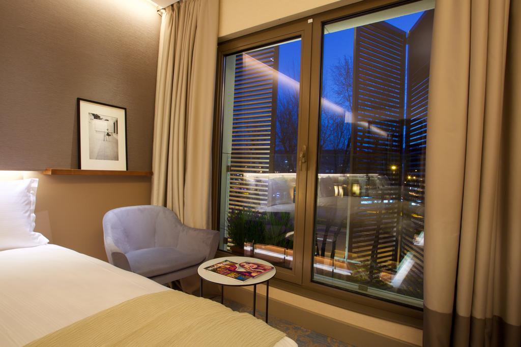 ดอสโซ่ ดอสซี่ โฮเต็ล แอน์ สปา ดาวน์ทาวน์ Hotel อิสตันบูล ภายนอก รูปภาพ The photo depicts a modern hotel room. On the left side, there is a neatly made bed with a light-colored blanket. Next to the bed, there's a small round table with some magazines on top. A cozy chair in a light shade is positioned nearby. The main fe